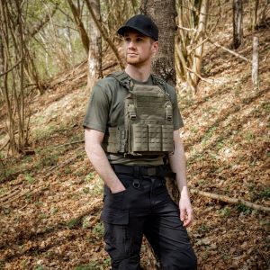 Homme en gilet tactique en Cordura 500D lasercut, chemise kaki, pantalon cargo noir, debout près d’un arbre en forêt.