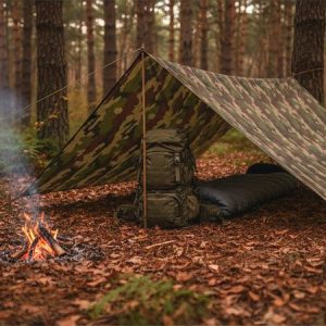 Bâche camoufle robuste installée comme abri de bivouac avec tapis de sol, feu de camp, forêt et sacs à l’arrière-plan.